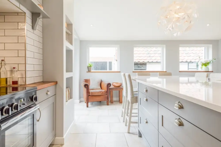 Bright classic kitchen with shaker-style grey cabinetry, white quartz island, built-in shelving, stainless steel range cooker, and a cozy dining nook with wooden chairs by three large windows.
