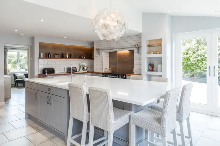 Open-plan classic kitchen with large grey shaker island, white quartz worktop, integrated breakfast bar seating, stainless steel range cooker, subway tile splashback, and pendant lighting near French doors to garden